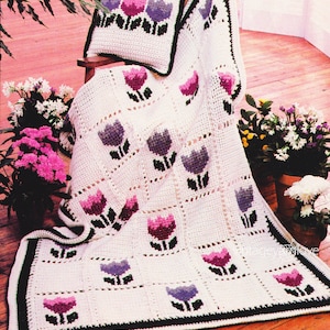 May include: A white crocheted throw blanket with a repeating pattern of purple, pink, and black tulips. The blanket is folded over a chair and has a matching pillow with the same tulip pattern.