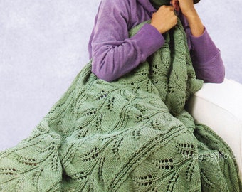 May include: A green crocheted blanket with a leafy pattern. The blanket is draped over a sofa and a woman is sitting on the sofa holding the blanket.
