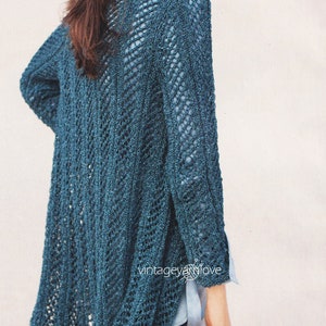 May include: A teal blue open knit cardigan with a lacy pattern. The cardigan is worn over a light blue shirt and dark grey pants.