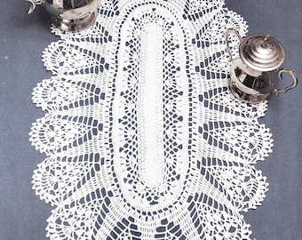 Può includere: Un runner da tavolo ovale in crochet bianco con un delicato motivo a pizzo. Il runner è disposto su una superficie blu con teiere e zuccheriere d'argento.