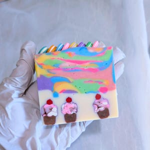 May include: A rectangular bar of handmade soap with a colorful, swirled design in shades of pink, yellow, blue, and purple. The soap features three cupcake designs with cherry toppings and a row of colorful candy pieces along the top.