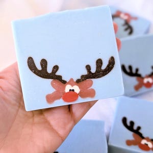 May include: Light blue square soap bars with a reindeer design. The reindeer has brown antlers, a red nose, and a light brown face. Several bars are visible, with one held in a hand.