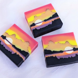 May include: Three square soaps with a sunset design. The soaps feature a black base with layers of purple, orange, yellow, and pink, and a white circular element. The soaps are arranged on a white surface.