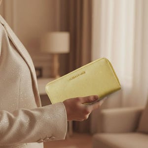 May include: A golden-yellow wallet with the text "ALREADY HAVE" in gold lettering. The wallet is rectangular and appears to be made of leather. The image shows the wallet being held by a person wearing a light-colored blazer.