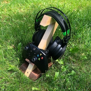 Op de afbeelding: Een houten standaard met een zwarte videogamecontroller en een zwarte koptelefoon met groene accenten. De standaard staat op groen gras.
