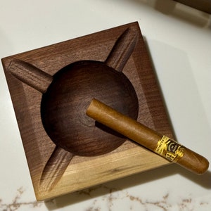 May include: A wooden cigar ashtray with a deep well and four slots. A brown cigar with a gold band is resting in the well. The band reads "COHIBA" and "18 de Diciembre".