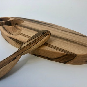 May include: A wooden serving tray with two small bowls and a wooden spreader. The tray is oval shaped and has a natural wood finish with dark brown streaks. The spreader has a curved handle.
