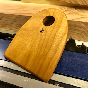 May include: A wooden scraper with a rounded shape and a hole in the center. The scraper has a natural wood finish and a black symbol on the surface.