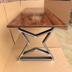 May include: A rustic wooden table top with a dark brown stain sits on a metal base with a starburst design. The table is made of reclaimed wood and has a natural, distressed look.