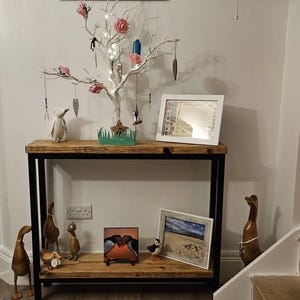May include: A wooden console table with a black metal frame, decorated with wooden ducks, framed pictures, and a white decorative tree with lights and ornaments. A penguin figurine is also on display. The table is against a white wall.