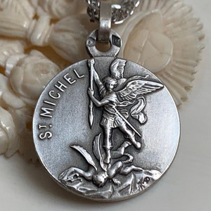 Op de afbeelding: Zilveren ketting met hanger met een afbeelding van Sint Michaël de Aartsengel die een demon verslaat. De hanger is rond en heeft de woorden "St. Michel" rond de rand gegraveerd.