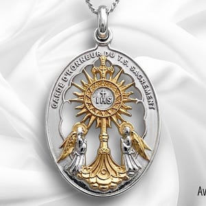 May include: Silver and gold-toned pendant necklace with an oval shape. The pendant features a sunburst design with the letters "IHS" at the center, flanked by two angel figures. The text "GARDE D'HONNEUR DU T.S. SACREMENT" is inscribed around the edge.