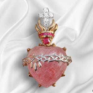 Può includere: Un ciondolo a forma di cuore con una pietra rosa testurizzata avvolta da una corona di spine argentata. Sopra il cuore, una fiamma dorata è decorata con gemme rosse e sormontata da una croce argentata incastonata con pietre trasparenti.
