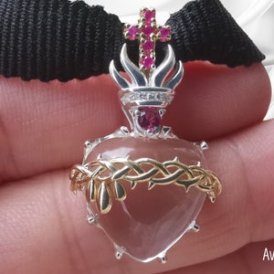 Peut inclure: Pendentif en forme de cœur avec un design argenté et doré. Le pendentif présente une couronne d'épines, une croix avec des pierres précieuses rouges et un rubis central. Il est attaché à un ruban noir.