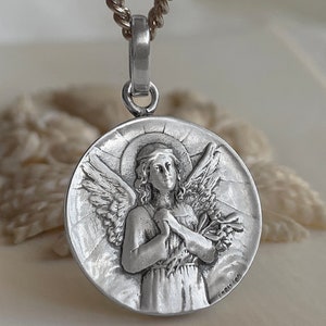 May include: Silver pendant necklace featuring a detailed relief of an angel with wings outstretched, holding a bouquet of flowers. The angel has a halo and is wearing a long robe. The pendant is round and has a textured surface.