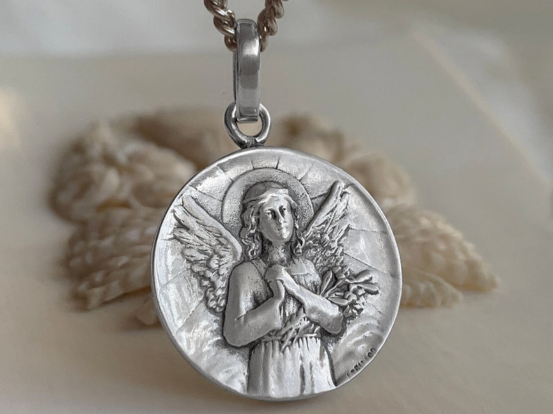 May include: Silver pendant necklace featuring a detailed relief of an angel with wings outstretched, holding a bouquet of flowers. The angel has a halo and is wearing a long robe. The pendant is round and has a textured surface.