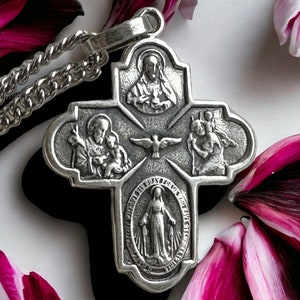 May include: A silver-colored pendant necklace featuring a cross design with religious figures and symbols. The pendant is suspended from a chain. The background includes a white surface and dark pink flower petals.