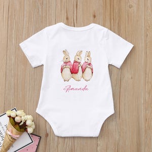 May include: White baby onesie with a pink and brown illustration of three bunnies wearing red coats. The onesie has the name "Amanda" printed in pink below the bunnies.