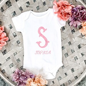 Pu&ograve; includere: Un body per neonati bianco a maniche corte con una grande lettera S rosa in stile acquerello stampata al centro. Sotto la lettera, il nome SOPHIA &egrave; stampato in un carattere serif rosa abbinato. Il capo presenta uno scollo rotondo.