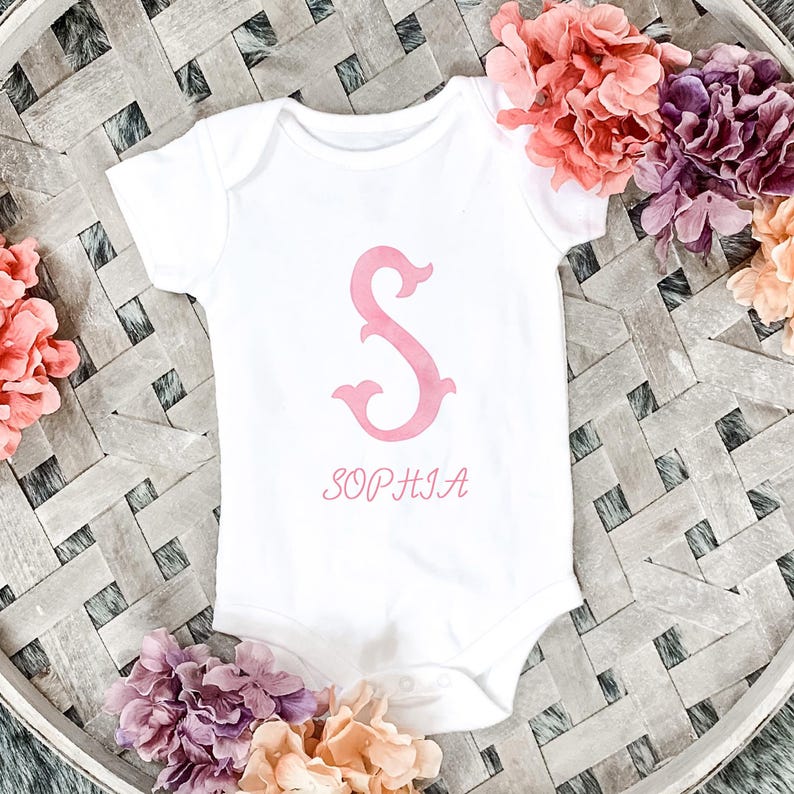 Pu&ograve; includere: Un body per neonati bianco a maniche corte con una grande lettera S rosa in stile acquerello stampata al centro. Sotto la lettera, il nome SOPHIA &egrave; stampato in un carattere serif rosa abbinato. Il capo presenta uno scollo rotondo.