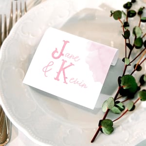 Pu&ograve; includere: Un segnaposto rettangolare bianco con testo rosa che recita Jane & Kevin. Il cartoncino presenta una delicata sfumatura ad acquerello rosa sul lato destro. La scritta combina lettere maiuscole graziate in grassetto e un font corsivo sottile.