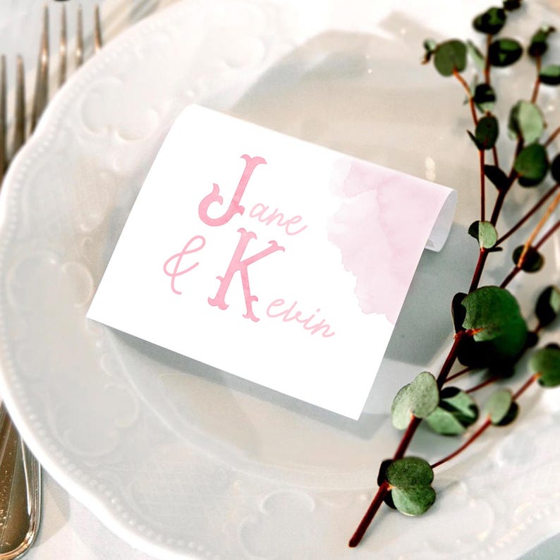 Pu&ograve; includere: Un segnaposto rettangolare bianco con testo rosa che recita Jane & Kevin. Il cartoncino presenta una delicata sfumatura ad acquerello rosa sul lato destro. La scritta combina lettere maiuscole graziate in grassetto e un font corsivo sottile.