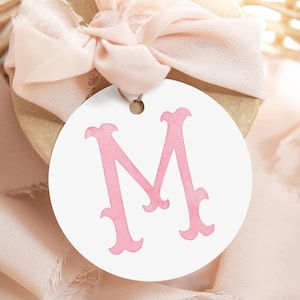 Pu&ograve; includere: Un'etichetta regalo circolare bianca con una grande lettera M stilizzata stampata in una tonalit&agrave; rosa tenue. L'etichetta presenta un piccolo foro nella parte superiore per l'aggancio ed &egrave; posizionata su uno sfondo in tessuto testurizzato con un nastro rosa pallido.