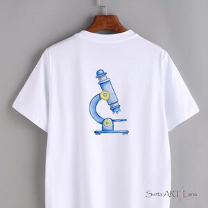 May include: White t-shirt featuring a blue and yellow watercolor illustration of a microscope. The design is centered on the back of the shirt. The shirt has short sleeves and a crew neck.