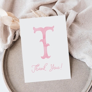 Pu&ograve; includere: Un biglietto d'auguri rettangolare bianco con una grande lettera T rosa in stile acquerello centrata in alto. Sotto la lettera, le parole Thank You! sono stampate in un carattere corsivo rosa abbinato nella parte inferiore del biglietto.