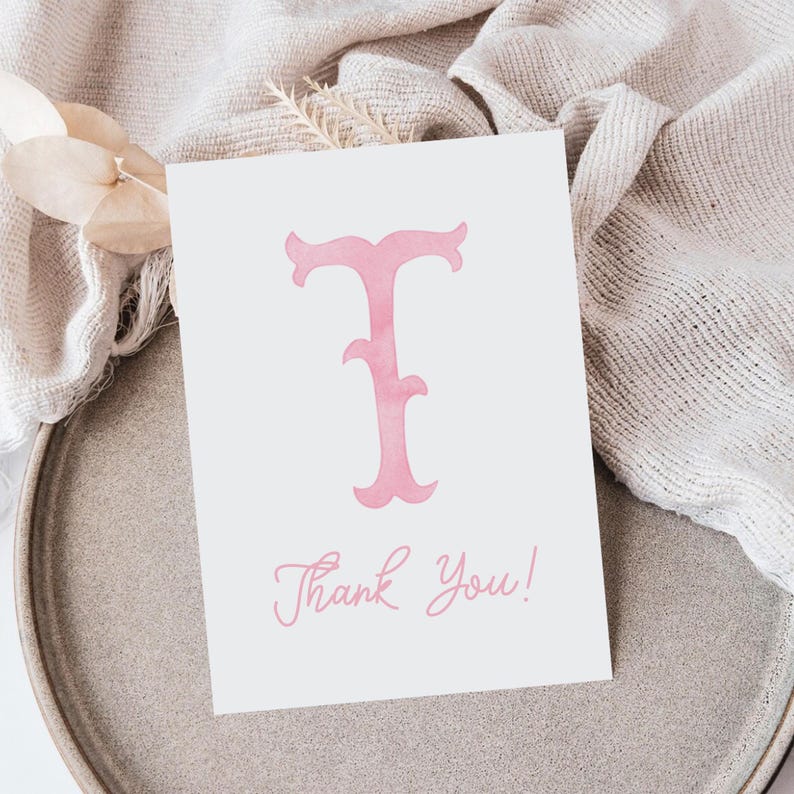Pu&ograve; includere: Un biglietto d'auguri rettangolare bianco con una grande lettera T rosa in stile acquerello centrata in alto. Sotto la lettera, le parole Thank You! sono stampate in un carattere corsivo rosa abbinato nella parte inferiore del biglietto.