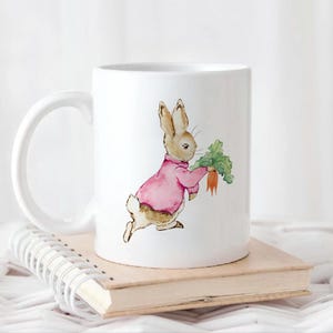 May include: White ceramic mug with a cartoon illustration of a brown rabbit wearing a pink sweater and holding a carrot. The rabbit is running to the right.