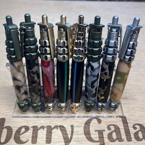 Puede incluir: Una colección de ocho bolígrafos hechos a mano exhibidos en un soporte acrílico transparente. Los bolígrafos presentan diseños únicos, incluyendo camuflaje y patrones veteados. Cada bolígrafo tiene un clip y una parte superior metálicos.