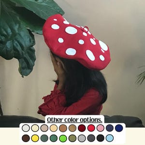 May include: A red mushroom-shaped hat with white polka dots. The hat is made of a soft material and has a small loop at the top. The image also shows a selection of other color options.