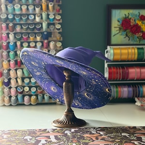 Peut inclure: Un chapeau de sorcière violet avec un motif céleste de lunes et d'étoiles dorées, présenté sur un présentoir en bois décoratif. Le chapeau a un large bord et une couronne pointue. En arrière-plan, des bobines de fil, des rubans et une broderie florale encadrée.