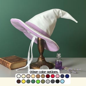 Pode incluir: Um chapéu de bruxa branco com aba e faixa lavanda, exibido em um suporte decorativo. A imagem inclui um livro antigo, uma pequena garrafa de vidro e raminhos de lavanda. Uma paleta de cores de outras opções de cores também é exibida.