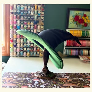 Pode incluir: Um chapéu de bruxa preto com uma aba verde é exibido em um suporte de madeira decorativo. O chapéu é o ponto focal, em um cenário de materiais de costura e um bordado floral.