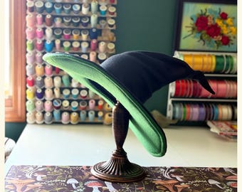 May include: A black witch hat with a green brim is displayed on a decorative wooden stand. The hat is the focal point, set against a backdrop of sewing supplies and a floral embroidery.