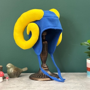 Puede incluir: Un sombrero azul con grandes cuernos de carnero amarillos, orejeras y una correa para la barbilla. El sombrero se exhibe en un soporte decorativo. El sombrero es probablemente para un niño o un disfraz. El sombrero está hecho de tela suave.