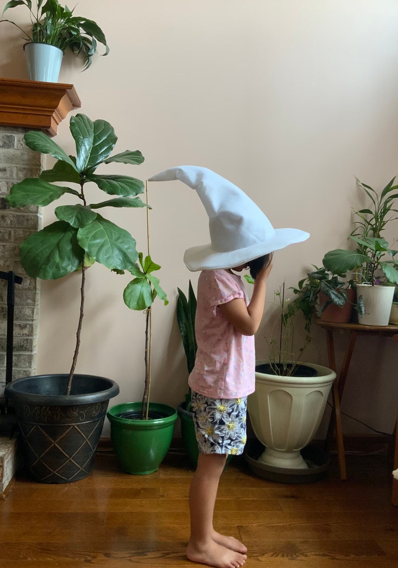 May include: A child wearing a white pointed hat with a wide brim, a pink shirt, and floral print shorts. The child is standing in a room with plants and a wooden floor.