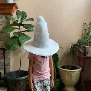 May include: A child wearing a white pointed hat with a wide brim. The hat is covering the child's face. The child is wearing a pink shirt and floral print shorts. The child is standing in front of a brick fireplace with plants in pots.