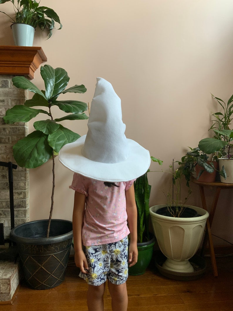 May include: A child wearing a white pointed hat with a wide brim. The hat is covering the child's face. The child is wearing a pink shirt and floral print shorts. The child is standing in front of a brick fireplace with plants in pots.