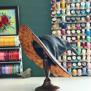 Puede incluir: Un sombrero de bruja gris oscuro con ala ancha, exhibido en un soporte decorativo. La parte inferior del ala presenta un vibrante patrón de hojas de otoño en tonos naranja y marrón. El fondo incluye carretes de hilo y cintas coloridas.