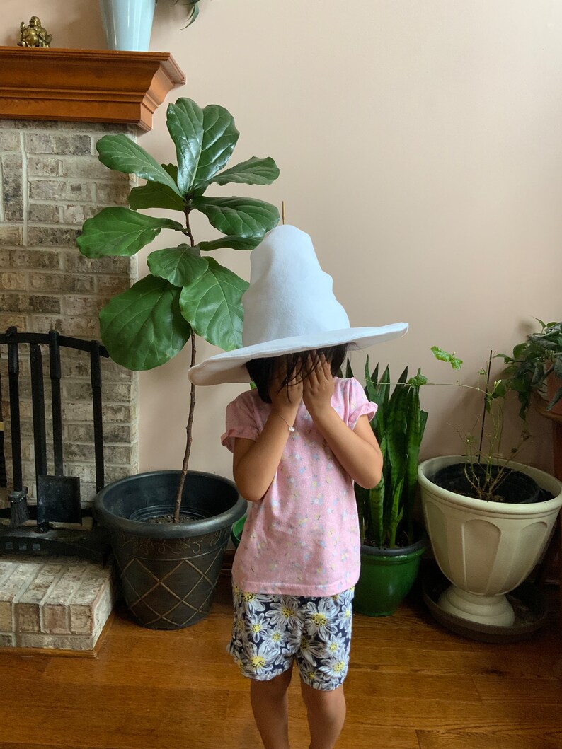 May include: A child wearing a white pointed hat, a pink shirt with a floral pattern, and floral print shorts. The child is standing in front of a fireplace and plants.