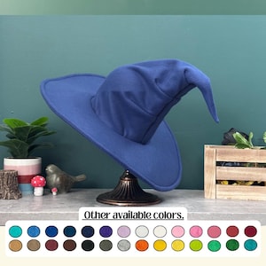 Puede incluir: Un sombrero de bruja azul marino con una corona puntiaguda y ala ancha, exhibido en un soporte decorativo. La imagen incluye el texto "Otros colores disponibles." y una paleta de colores variados.