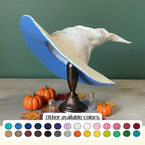 May include: A beige witch hat with a light blue brim, displayed on a decorative stand. The hat is surrounded by small orange pumpkins, autumn leaves, and miniature glass bottles. The text "Other available colors" is at the bottom.