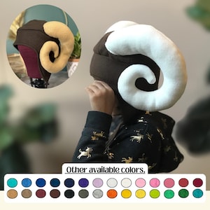 May include: A brown hat with large, curved, cream-colored horns. The hat has a dark brown base and is shown on a child. The image also displays a color palette of available hat colors, including teal, navy, and pink.