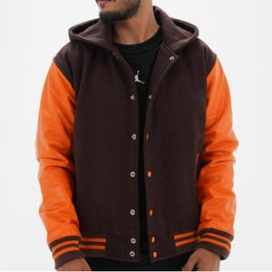 Puede incluir: Chaqueta universitaria con capucha marrón con mangas de cuero naranja. La chaqueta tiene una botonadura y rayas naranjas en los puños y el dobladillo. La capucha es marrón, a juego con el cuerpo de la chaqueta.