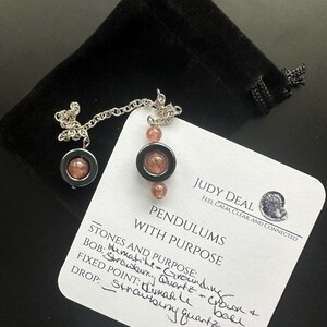 May include: A pendulum with a silver chain and two strawberry quartz beads. The pendulum is accompanied by a black velvet pouch and a white card with handwritten details and the text "PENDULUMS WITH PURPOSE".