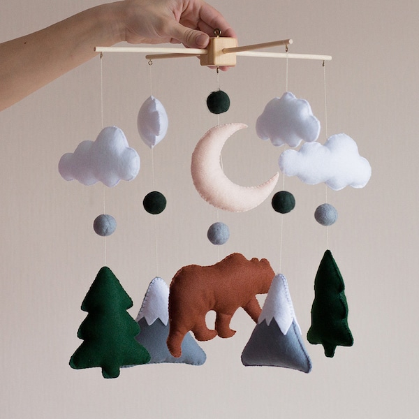 May include: A felt baby mobile with a brown bear, green trees, gray mountains, white clouds, a pink crescent moon, and gray and green balls.