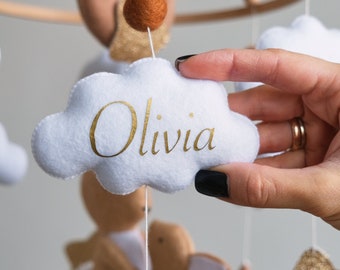 Puede incluir: Móvil para bebé con un aro de madera, con nubes de fieltro blanco, una luna de fieltro marrón y un conejito de fieltro. La nube en primer plano tiene el nombre "Olivia" en letras doradas. Una mano sostiene la nube.